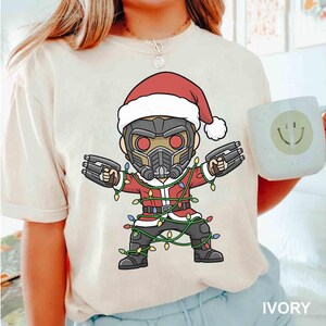 May include: Ivory-colored t-shirt featuring a cartoon character in a Santa hat and suit, holding blasters. The character is wrapped in Christmas lights. A white mug with a smiley face is held in the background. The shirt is labeled "IVORY".