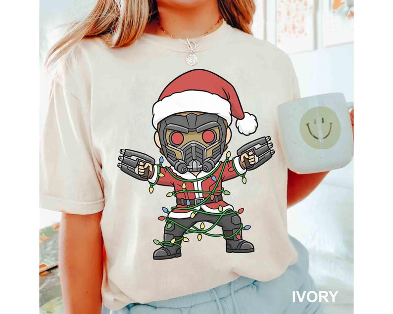 May include: Ivory-colored t-shirt featuring a cartoon character in a Santa hat and suit, holding blasters. The character is wrapped in Christmas lights. A white mug with a smiley face is held in the background. The shirt is labeled "IVORY".