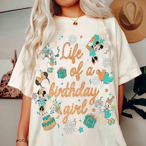 May include: Cream-colored t-shirt with a colorful graphic featuring Disney characters and the text "Life of a birthday girl." The design includes Minnie Mouse, Daisy Duck, Mickey Mouse, and a birthday cake, with fireworks and gift boxes.