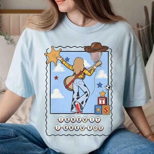 May include: A light blue t-shirt featuring a graphic print of a person with long hair wearing a yellow plaid shirt and cow-print pants, holding a guitar and a brown cowboy hat. The design includes a sheriff star, blocks with the letters T and S, and the text I KNEW IT I KNEW YOU.