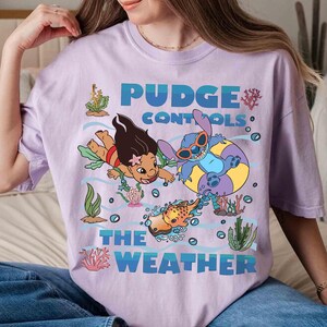 May include: A light purple t-shirt featuring a graphic of characters from the movie Lilo & Stitch. The text "Pudge Controls The Weather" is in blue. The design includes cartoon characters, fish, and aquatic plants.