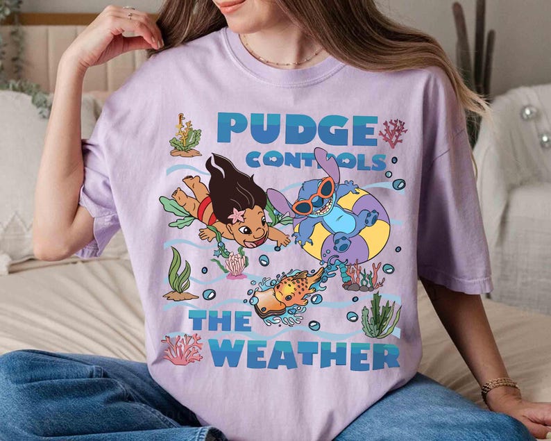 May include: A light purple t-shirt featuring a graphic of characters from the movie Lilo & Stitch. The text "Pudge Controls The Weather" is in blue. The design includes cartoon characters, fish, and aquatic plants.