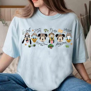 May include: Light blue t-shirt featuring cartoon characters Goofy, Donald Duck, Minnie Mouse, Pluto, Mickey Mouse, and Daisy Duck. Each character is wearing a safari hat. The shirt has the characters' names written above them in a handwritten style.