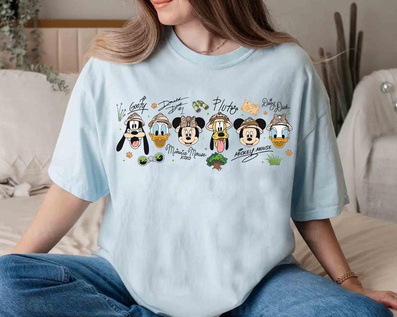 May include: Light blue t-shirt featuring cartoon characters Goofy, Donald Duck, Minnie Mouse, Pluto, Mickey Mouse, and Daisy Duck. Each character is wearing a safari hat. The shirt has the characters' names written above them in a handwritten style.