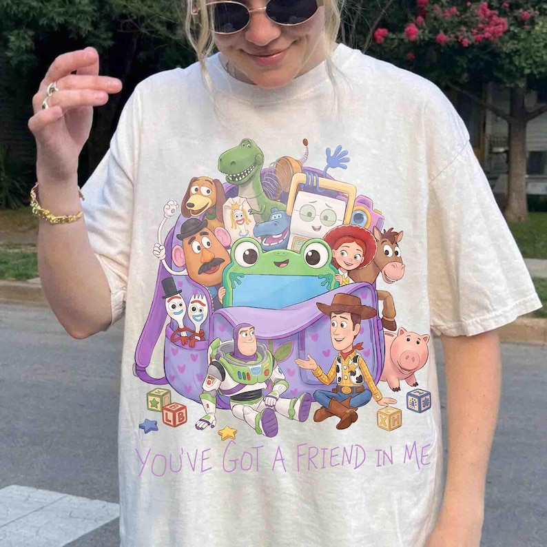 May include: A cream-colored short-sleeve t-shirt featuring a graphic print of various Toy Story characters emerging from a purple backpack. The text "YOU'VE GOT A FRIEND IN ME" is printed in purple below the graphic, which includes Buzz Lightyear, Woody, and other animated figures.