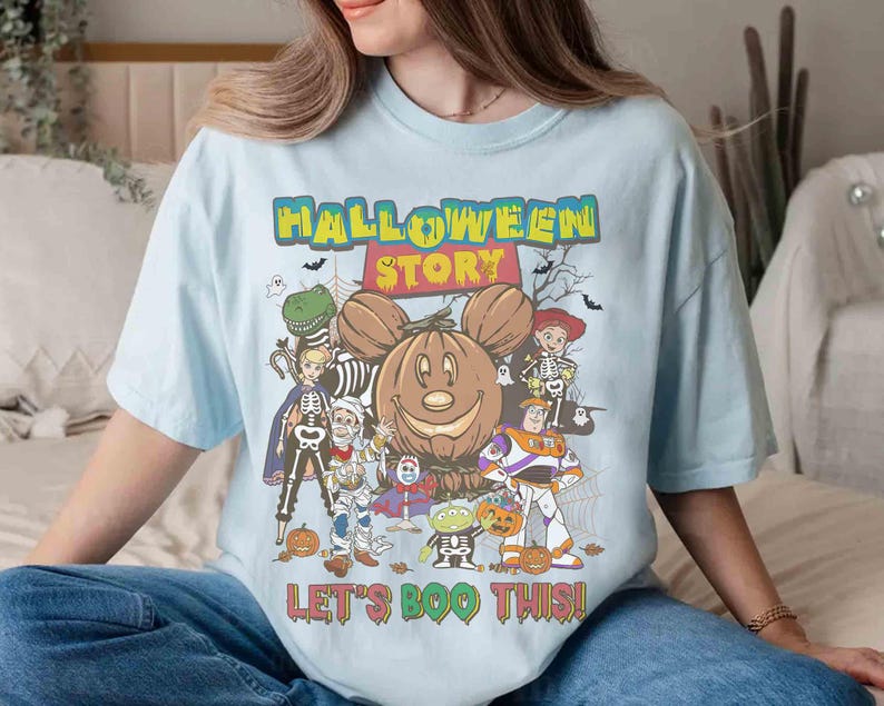 Puede incluir: Camiseta azul claro con un dise&ntilde;o tem&aacute;tico de Halloween. El dise&ntilde;o presenta un Mickey Mouse con forma de calabaza, personajes de dibujos animados con disfraces de Halloween y el texto "HALLOWEEN STORY" y "LET'S BOO THIS!"