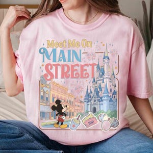 May include: Pink t-shirt with a graphic featuring Mickey Mouse, a castle, and the text "Meet Me On Main Street." The design includes fireworks and other park-themed elements. The shirt is a light pink color.
