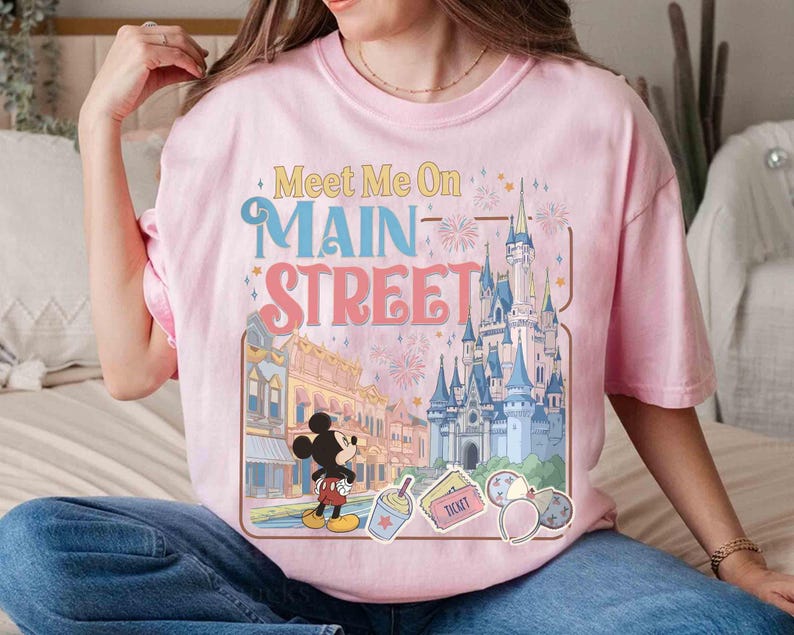 May include: Pink t-shirt with a graphic featuring Mickey Mouse, a castle, and the text "Meet Me On Main Street." The design includes fireworks and other park-themed elements. The shirt is a light pink color.
