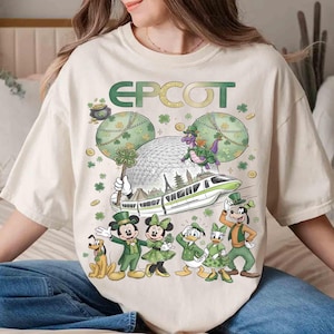 May include: Cream-coloured t-shirt with a St. Patrick's Day EPCOT design. The graphic features Mickey and Minnie Mouse, Donald and Daisy Duck, Goofy, and Pluto in green outfits, a monorail, and the EPCOT sphere.