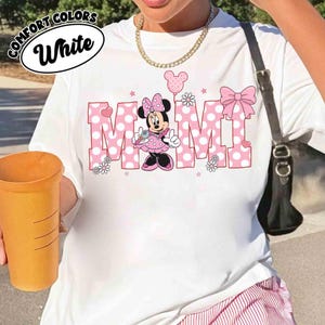 Puede incluir: Camiseta blanca con la palabra "MOM" en letras rosas de lunares, con un gr&aacute;fico de Minnie Mouse. La camiseta incluye un lazo, flores y estrellas. Las palabras "COMFORT COLORS" y "White" est&aacute;n en un c&iacute;rculo en la parte superior.