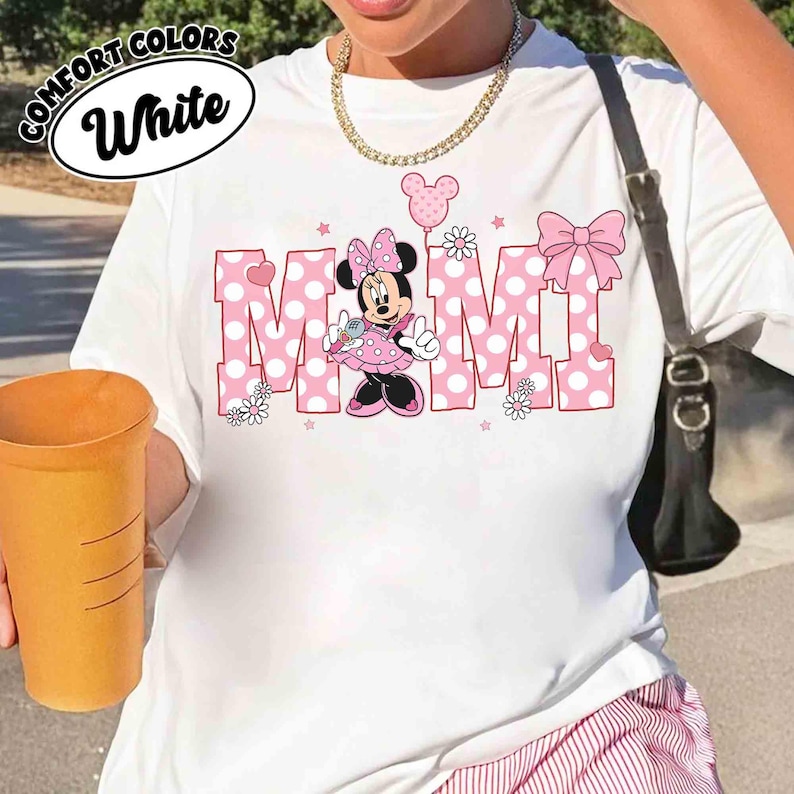 Puede incluir: Camiseta blanca con la palabra "MOM" en letras rosas de lunares, con un gr&aacute;fico de Minnie Mouse. La camiseta incluye un lazo, flores y estrellas. Las palabras "COMFORT COLORS" y "White" est&aacute;n en un c&iacute;rculo en la parte superior.