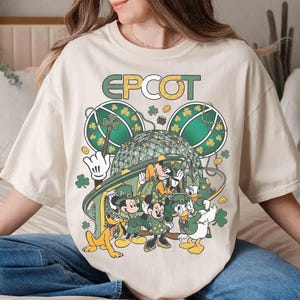 Puede incluir: Camiseta color crema con un gráfico de personajes de Disney vestidos de verde para el Día de San Patricio. El diseño incluye Mickey y Minnie Mouse, Donald y Daisy Duck y Pluto, con la palabra "EPCOT" arriba.