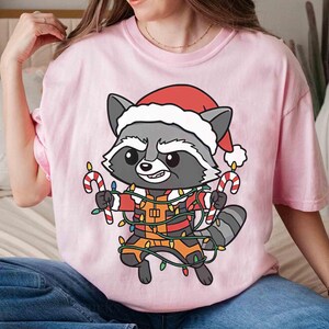 May include: A light pink t-shirt featuring a cartoon raccoon wearing a Santa hat and holding candy canes. The raccoon is wrapped in Christmas lights. The shirt is a festive holiday design.