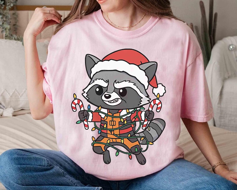 May include: A light pink t-shirt featuring a cartoon raccoon wearing a Santa hat and holding candy canes. The raccoon is wrapped in Christmas lights. The shirt is a festive holiday design.