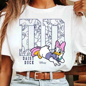 May include: White t-shirt featuring a graphic of Daisy Duck in a relaxed pose. The number "10" is printed in navy blue with a Daisy Duck pattern inside. The words "DAISY DUCK" and the Disney logo are also printed on the shirt.