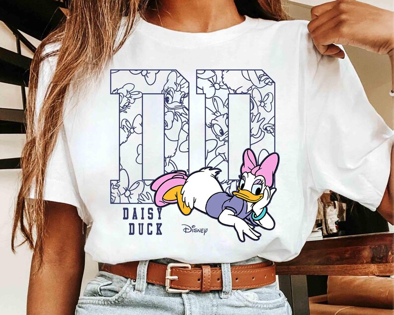 May include: White t-shirt featuring a graphic of Daisy Duck in a relaxed pose. The number "10" is printed in navy blue with a Daisy Duck pattern inside. The words "DAISY DUCK" and the Disney logo are also printed on the shirt.
