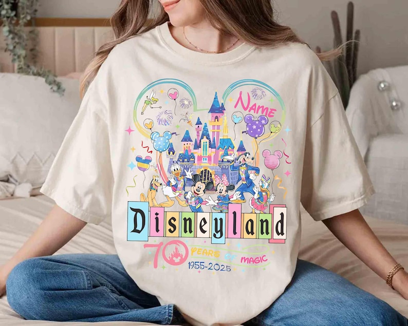 May include: Cream-colored t-shirt featuring a colorful Disneyland graphic. The design includes a castle, Mickey Mouse, Minnie Mouse, Donald Duck, and other characters. The text reads "Disneyland" and "70 Years of Magic 1955-2025".
