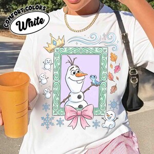 May include: A white t-shirt featuring a graphic print of a snowman and a small blue lizard inside a decorative green frame. The design includes a gold crown, a pink bow, small snowmen, snowflakes, and autumn leaves surrounding the frame.