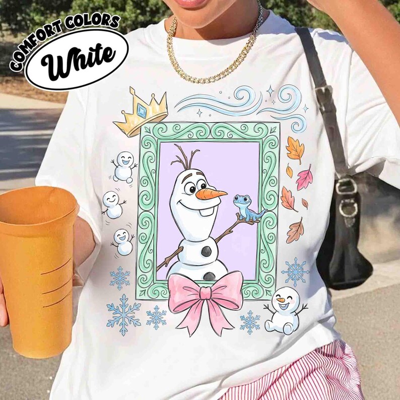 May include: A white t-shirt featuring a graphic print of a snowman and a small blue lizard inside a decorative green frame. The design includes a gold crown, a pink bow, small snowmen, snowflakes, and autumn leaves surrounding the frame.