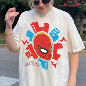 Puede incluir: Una camiseta de manga corta color crema con un gr&aacute;fico central de la m&aacute;scara de Spider-Man rodeada por siluetas de edificios rojos y azules y formas de rayos. El texto dice Marvel Studios Spider-Man Brand New Day.