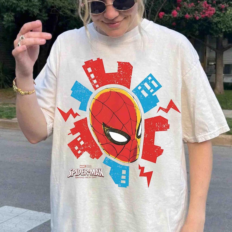 Puede incluir: Una camiseta de manga corta color crema con un gr&aacute;fico central de la m&aacute;scara de Spider-Man rodeada por siluetas de edificios rojos y azules y formas de rayos. El texto dice Marvel Studios Spider-Man Brand New Day.