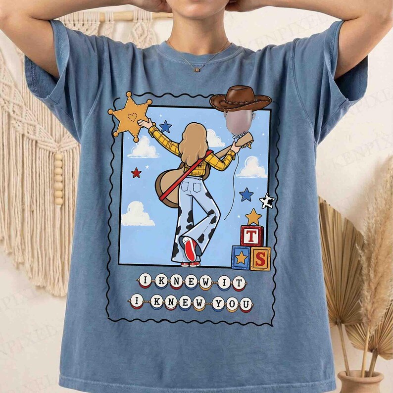 May include: A blue t-shirt featuring a graphic of a person with long hair wearing a yellow plaid shirt and cow-print pants, holding a guitar. The design includes a cowboy hat, a sheriff star, building blocks with letters T and S, and the text I KNEW IT I KNEW YOU.