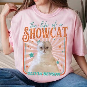 Może przedstawiać: R&oacute;żowa koszulka z grafiką białego i szarego kota z napisem "the life of a SHOWCAT" w kolorze pomarańczowym i turkusowym. Grafika kota umieszczona jest na tle retro z gwiazdami i nazwą "OLIVIA-BENSON" poniżej.