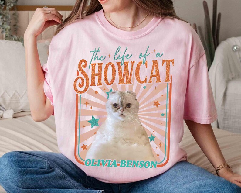 Może przedstawiać: R&oacute;żowa koszulka z grafiką białego i szarego kota z napisem "the life of a SHOWCAT" w kolorze pomarańczowym i turkusowym. Grafika kota umieszczona jest na tle retro z gwiazdami i nazwą "OLIVIA-BENSON" poniżej.