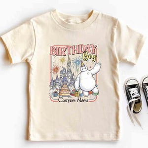 May include: Cream-colored t-shirt featuring a colorful graphic with the text "BIRTHDAY BOY" and "Custom Name". The graphic includes a cartoon character, a castle, a birthday cake, and presents. Black and white sneakers are next to the shirt.