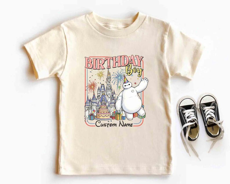 May include: Cream-colored t-shirt featuring a colorful graphic with the text "BIRTHDAY BOY" and "Custom Name". The graphic includes a cartoon character, a castle, a birthday cake, and presents. Black and white sneakers are next to the shirt.