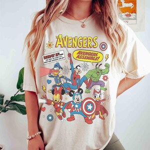 May include: A cream-coloured t-shirt with a vibrant graphic of The Avengers characters, including Mickey Mouse as Captain America, Iron Man Minnie, and Donald Duck as a superhero. Text includes "The Avengers" and "Avengers Assemble!"