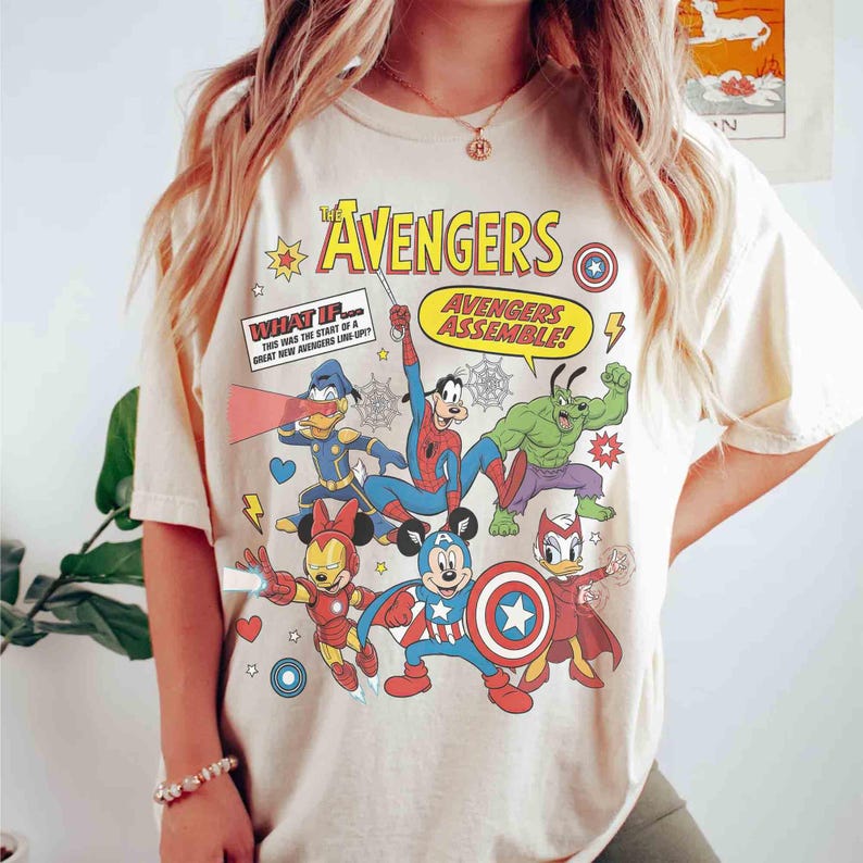 May include: A cream-coloured t-shirt with a vibrant graphic of The Avengers characters, including Mickey Mouse as Captain America, Iron Man Minnie, and Donald Duck as a superhero. Text includes "The Avengers" and "Avengers Assemble!"