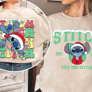 May include: Beige sweatshirt featuring the word "STITCH" in colorful, retro-style lettering with a cartoon Stitch character wearing a Santa hat. The design includes a festive holiday theme with lights and a playful aesthetic.