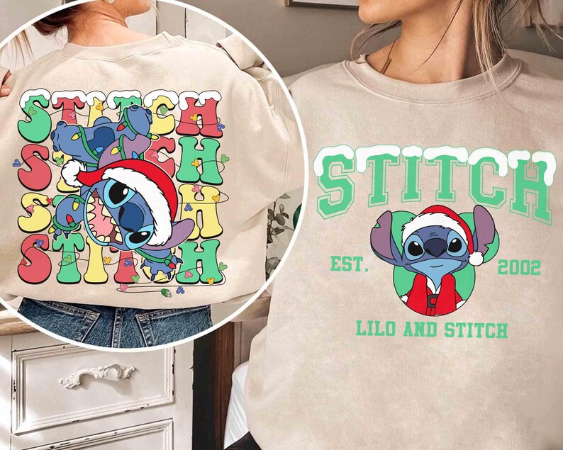 May include: Beige sweatshirt featuring the word "STITCH" in colorful, retro-style lettering with a cartoon Stitch character wearing a Santa hat. The design includes a festive holiday theme with lights and a playful aesthetic.