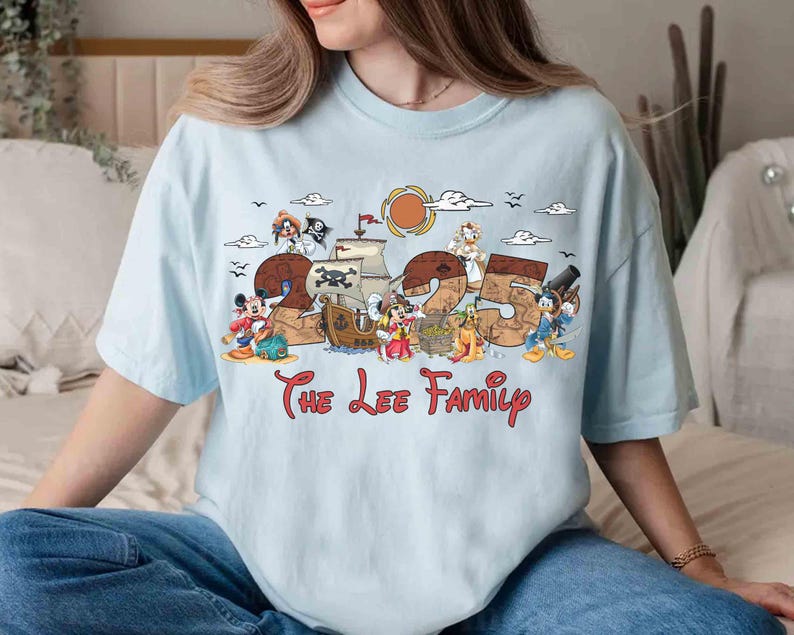 May include: Pale blue t-shirt with a vibrant graphic design incorporating the number "25" and cartoon characters like Mickey Mouse and Donald Duck. The words "The Lee Family" are printed beneath the design.