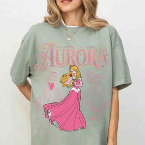 Op de afbeelding: Saliegroen t-shirt met het woord "Aurora" in roze letters en bloemmotieven. Een cartoonprinses in een roze jurk met een roos eronder. Vage prinsessenomtrekken staan ook op het shirt.