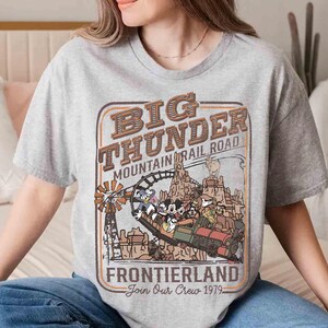 May include: A gray t-shirt featuring a vintage-style graphic for Big Thunder Mountain Railroad. The design includes a train with cartoon characters, the text "Frontierland," and "Join Our Crew 1979."