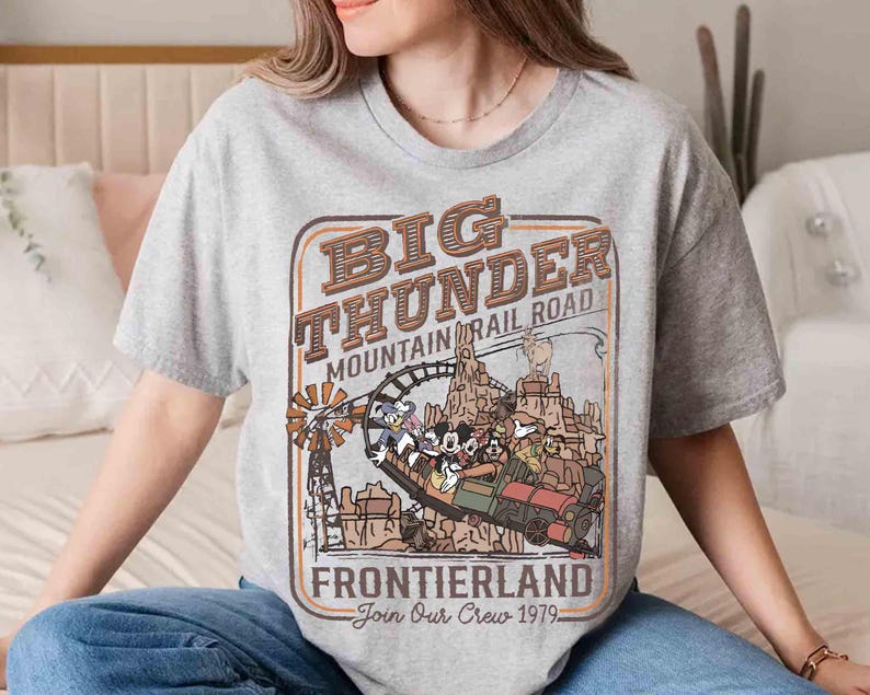May include: A gray t-shirt featuring a vintage-style graphic for Big Thunder Mountain Railroad. The design includes a train with cartoon characters, the text "Frontierland," and "Join Our Crew 1979."