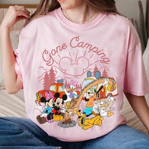 Pu&ograve; includere: T-shirt rosa chiaro con una grafica di personaggi Disney in campeggio. Il design include Topolino e Minnie, Paperino, Pippo e Paperina attorno a un fal&ograve;, con la scritta "Gone Camping".
