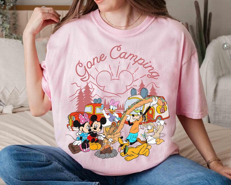 Pu&ograve; includere: T-shirt rosa chiaro con una grafica di personaggi Disney in campeggio. Il design include Topolino e Minnie, Paperino, Pippo e Paperina attorno a un fal&ograve;, con la scritta "Gone Camping".