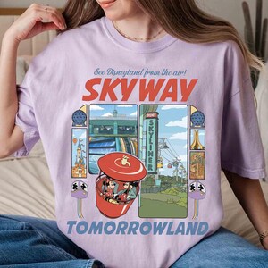 Puede incluir: Una camiseta lila con un gr&aacute;fico del paseo Skyway de Disneyland. El dise&ntilde;o incluye las palabras "SKYWAY" y "TOMORROWLAND", junto con ilustraciones del paseo e iconos del parque. La camiseta es de corte holgado.