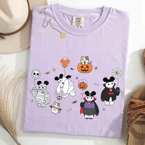 May include: A lilac Comfort Colors t-shirt featuring a Halloween-themed graphic. The design includes cartoon characters dressed as a mummy, vampire, and Baymax, along with a cat in a pumpkin, a spiderweb, and other Halloween elements.
