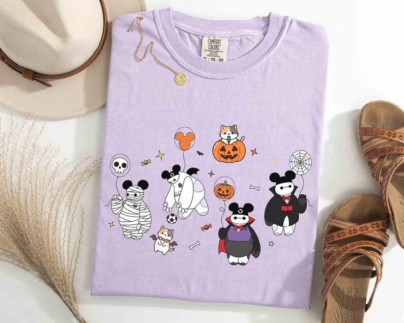 May include: A lilac Comfort Colors t-shirt featuring a Halloween-themed graphic. The design includes cartoon characters dressed as a mummy, vampire, and Baymax, along with a cat in a pumpkin, a spiderweb, and other Halloween elements.