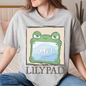 May include: A heather gray t-shirt featuring a cartoon frog holding a sign that says "Hi! Let's play!" in a blue and white design. The word "LILYPAD" is printed in a brown font below the frog illustration.