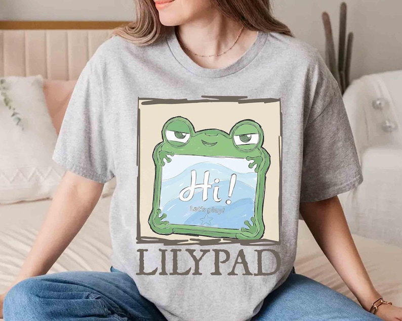 May include: A heather gray t-shirt featuring a cartoon frog holding a sign that says "Hi! Let's play!" in a blue and white design. The word "LILYPAD" is printed in a brown font below the frog illustration.