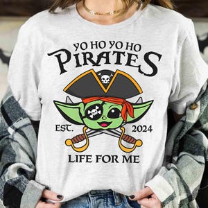 May include: A white t-shirt with a green alien wearing a pirate hat and holding crossed swords. The text on the shirt reads "Yo Ho Yo Ho Pirates Est. 2024 Life For Me".