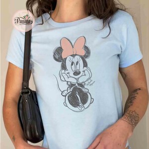 May include: A light blue t-shirt with a black and white sketch of Minnie Mouse on the front. Minnie Mouse is wearing a red bow and has a playful expression.