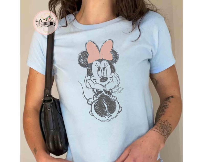 May include: A light blue t-shirt with a black and white sketch of Minnie Mouse on the front. Minnie Mouse is wearing a red bow and has a playful expression.