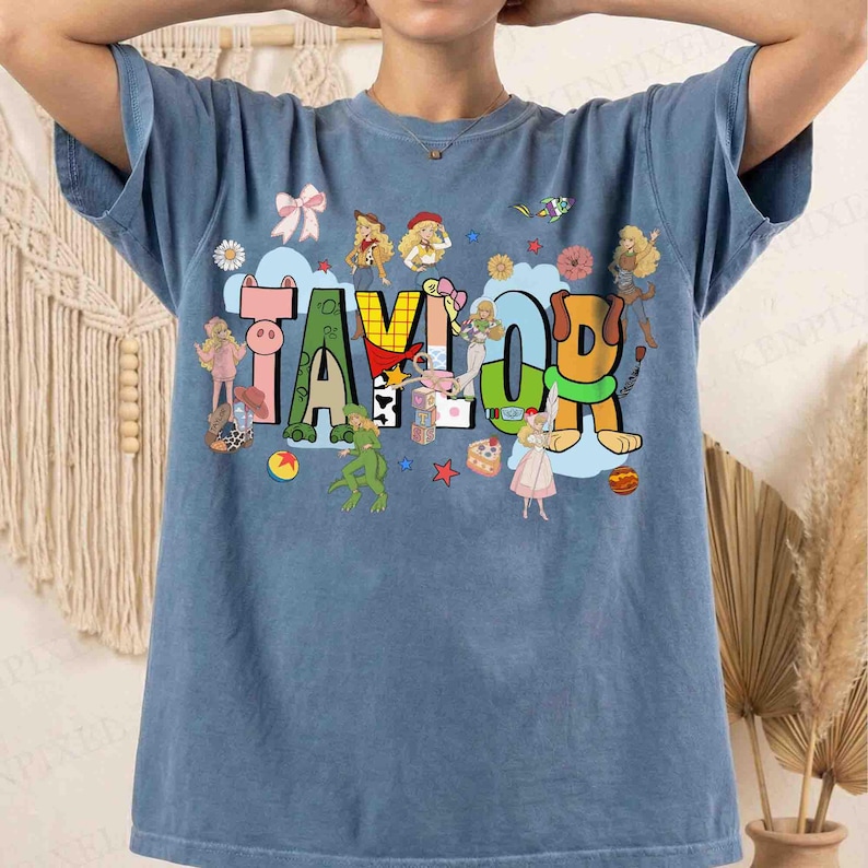 May include: A blue short-sleeve t-shirt featuring the name TAYLOR in large, colorful, block-style letters. The design includes various cartoon characters, a pink bow, a small cake, and stars scattered around the text on a light blue background.