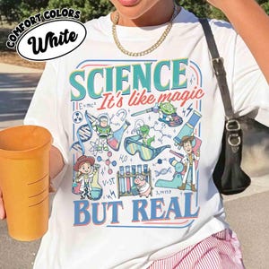 Pu&ograve; includere: T-shirt bianca con grafica colorata con la parola "SCIENCE" e la frase "It's like magic BUT REAL". Il design include personaggi dei cartoni animati e illustrazioni scientifiche. La maglietta ha anche le parole "COMFORT COLORS" e "White".